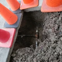 Excavated hole beside a concrete slab with a water pipe exposed and orange traffic cones around it
